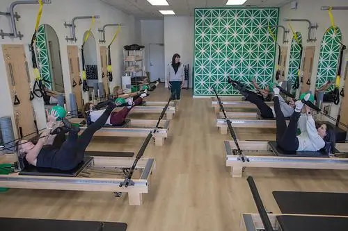 Women perform core exercises with green balls on reformers during a group Pilates class.
