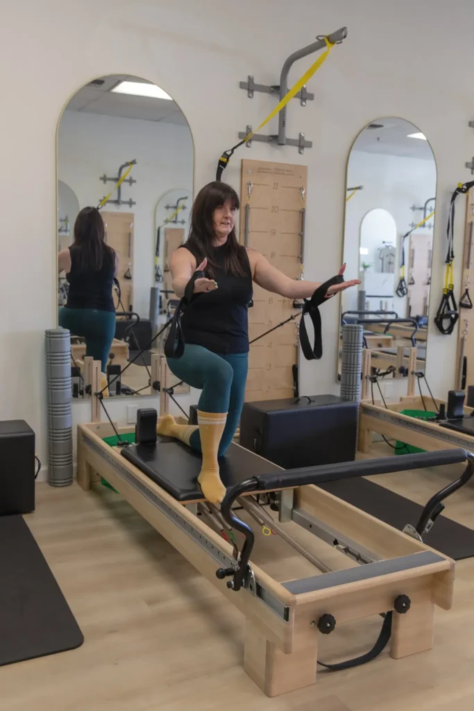Woman kneeling on a Pilates reformer performing an arm exercise with straps.